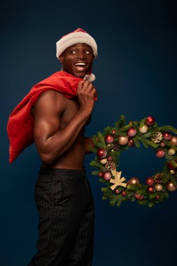 Gömleksiz ve neşeli Afro-Amerikan erkeği elinde şenlikli bir çelenk ve koyu mavi bir Noel çantası tutuyor.