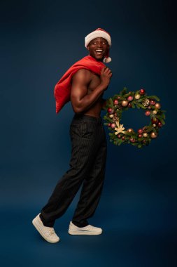 Neşeli ve gömleksiz Afro-Amerikalı adam elinde Noel çantası ve koyu mavi çelenkle yürüyor.