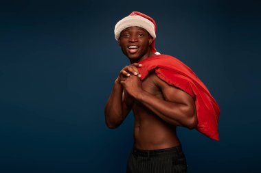 Noel Baba şapkalı, heyecanlı ve güçlü Afrikalı Amerikalı adam elinde Noel çantası ve koyu mavi üzerinde gülümsüyor.