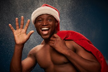 cheerful shirtless african american man with santa bag waving hand on dark blue snowy  backdrop clipart