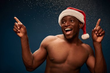 excited athletic african american man in santa hat pointing away with finger on dark snowy backdrop clipart