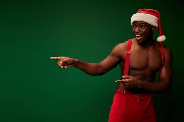 Noel Baba şapkalı, kırmızı pantolonlu, gülümseyen ve yeşili işaret eden üstsüz Afrikalı Amerikalı.