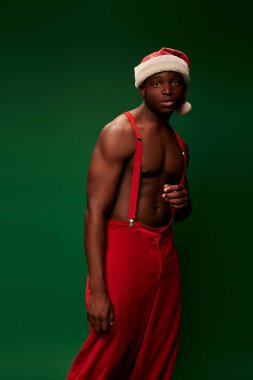 Gömleksiz, kaslı, Noel Baba şapkalı ve yeşil pantolonlu, düşünceli, Afro-Amerikalı bir adam.