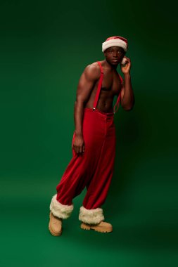 Düşünceli ve sportif, Noel Baba şapkalı ve kırmızı pantolonlu Afro-Amerikalı yeşil kameraya bakıyor.