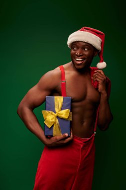 Neşeli atletik Afrikalı Amerikalı Noel Baba şapkalı adam hediyesini ve Noel çantasını yeşil üzerinde taşıyor.