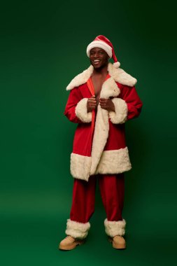 Geleneksel Noel Baba kostümlü neşeli Afro-Amerikalı adam yeşil fonda kameraya bakıyor.