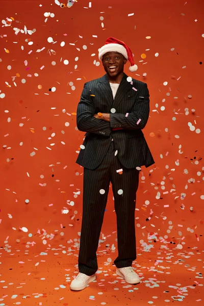 excited african american man in black suit and santa hat smiling under confetti on red backdrop