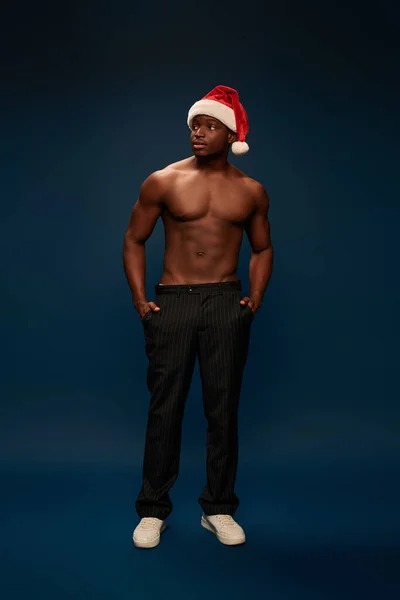 strong african american man in santa hat holding hands in pockets and looking away on dark blue