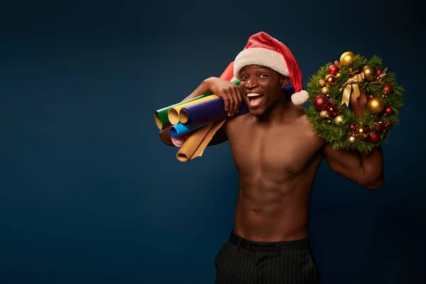 excited strong african american man in santa hat with wallpapers and christmas wreath on dark blue