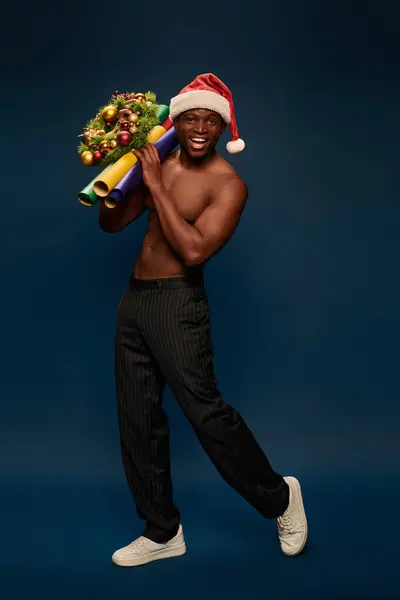 cheerful strong african american man in santa hat holding christmas decor on navy blue backdrop
