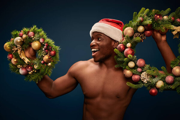 возбужденный безрубашечный африканский американец в Санта-Хапе, стоящий с рождественскими венками на темно-синем