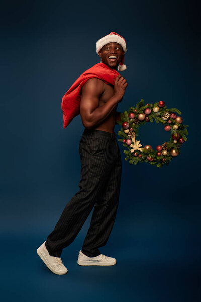 joyful and shirtless african american man walking with christmas bag and festive wreath on dark blue