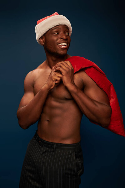 cheerful african american man with shirtless torso and christmas bag looking away on navy blue