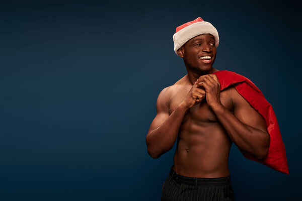 smiling african american man with muscular torso and christmas bag looking away on dark blue