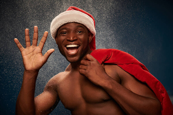 cheerful shirtless african american man with santa bag waving hand on dark blue snowy  backdrop