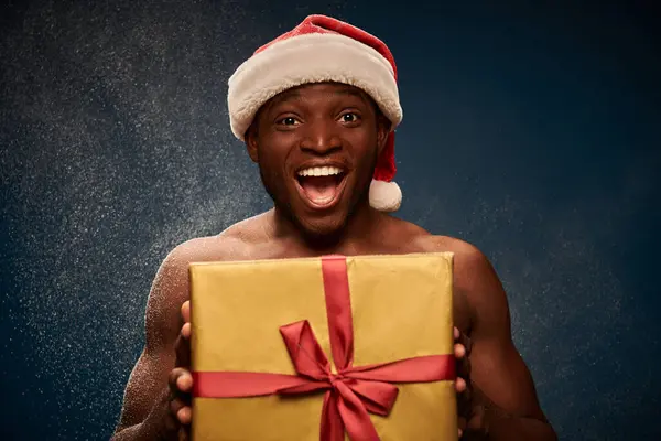 stock image thrilled sportive african american man in santa hat with shiny gift box on dark blue snowy backdrop