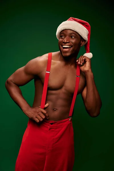 Stock image joyful shirtless african american man in santa hat and red pants looking away on green backdrop