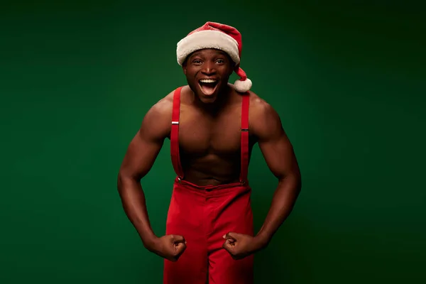 stock image energized and shirtless african american man in christmas hat and pants showing muscles on green