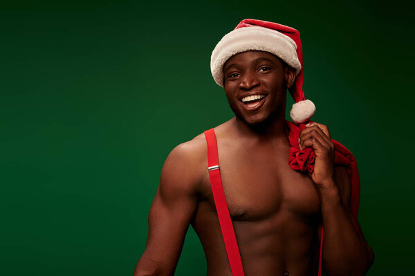 muscular african american man in santa hat holding christmas bags and looking at camera on green