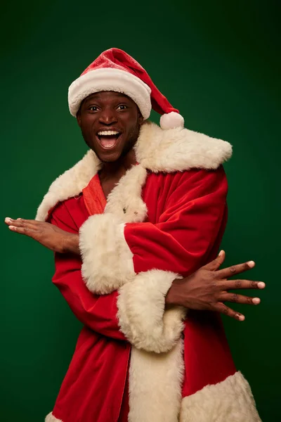 stock image overjoyed african american man in santa claus costume gesturing and looking at camera on green