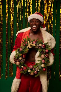 Mutlu Afro-Amerikan Noel Baba kostümlü adam, yeşil çelengin yanında Noel çelengi taşıyor.
