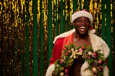 Noel Baba kostümlü gülen Afrikalı Amerikalı adam yeşil dekorun yanında Noel çelengi takıyor.