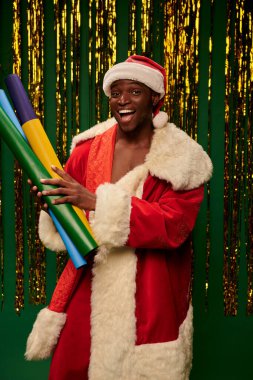 Noel Baba kostümü giymiş heyecanlı Afrikalı Amerikalı adam. Renkli duvar kağıtları ile yeşil arka planda süslemeli.