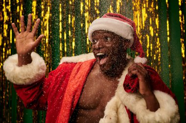 Noel Baba kostümü giymiş Afro-Amerikan adam. Elinde Noel çantası, karlı ve parlak arka planda el sallıyor.