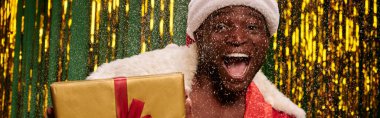 Karlı ve parlak stüdyoda hediye kutusuyla Noel Baba kostümü giymiş afili Afro-Amerikan bir adam.