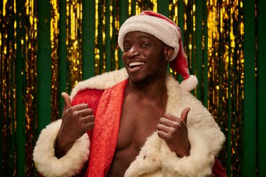 hot dark skinned guy in santa costume on shirtless body showing thumbs up on christmas backdrop clipart