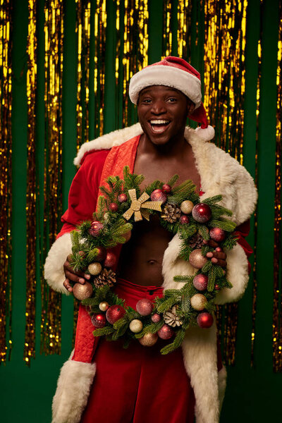 довольный африканский американец в костюме Санты с рождественским венком рядом с золотой мишурой на зеленом