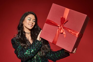 Yeşil ceketli mutlu Asyalı kadın kırmızı arka planda hediyesini tutuyor. Mutlu Noeller.