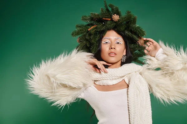 holiday spirit, beautiful asian woman with white makeup and trendy outfit posing in wreath on green