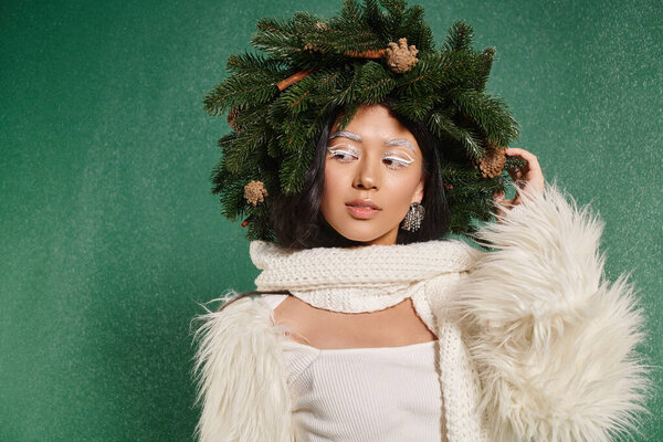 winter fashion, dreamy asian woman with natural wreath posing in white clothes under falling snow