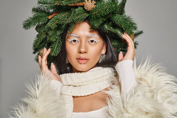 winter trends, attractive woman with white eye makeup and beads on face posing in wreath on grey