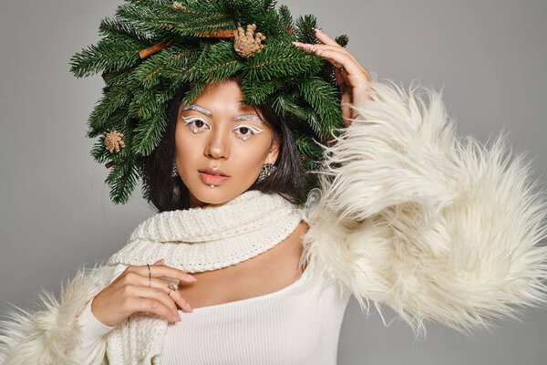 holiday style, charming woman with white eye makeup and beads on face posing in wreath on grey