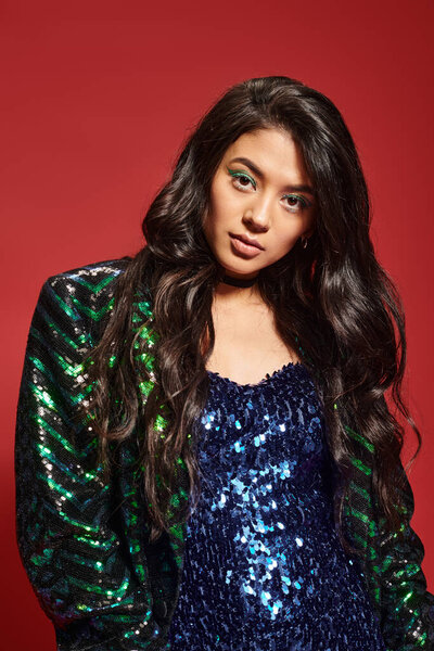 attractive asian woman in green dress and jacket with sequins looking at camera on red backdrop