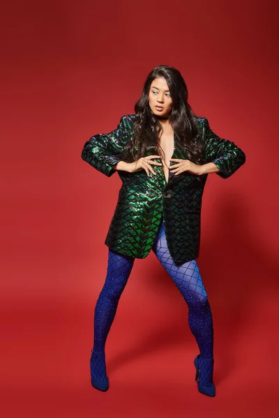 beautiful asian woman in green jacket with sequins and blue pantyhose posing on red background