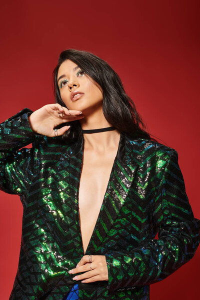 dreamy asian woman in trendy green jacket with sequins looking up and posing on red background