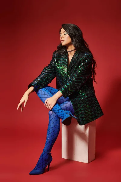 trendy asian woman in green jacket with sequins and blue pantyhose sitting on red backdrop