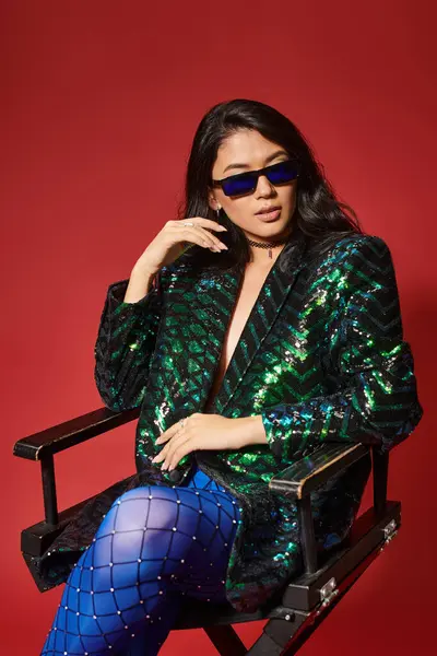 Stock image pretty asian woman in sunglasses and green jacket with sequins sitting on chair on red backdrop