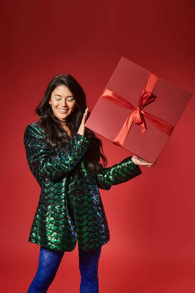 stock image happy asian woman in jacket with sequins and blue pantyhose holding present on red backdrop