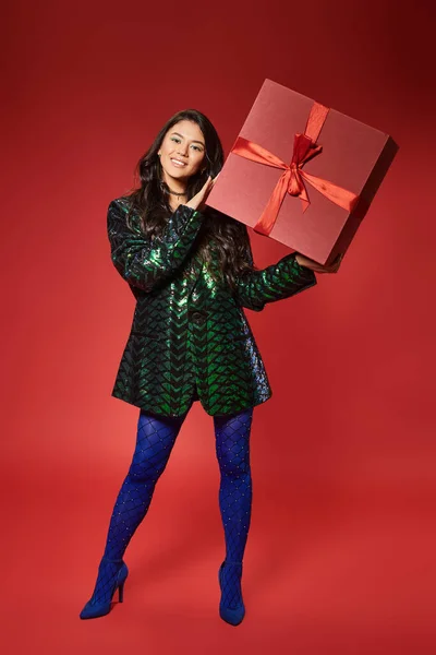 happy asian woman in green jacket with sequins holding gift box on red backdrop, Merry Christmas