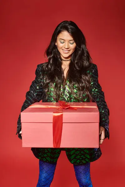 joyful asian woman in green jacket with sequins looking at gift box on red backdrop, Merry Christmas
