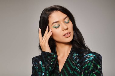 attractive asian woman in green jacket with sequins and bold eye makeup posing on grey backdrop clipart
