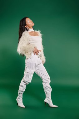 Beyaz kış kıyafetleri ve yeşil arka planda poz veren topuklu ayakkabıları olan Asyalı güzel bir kadın.