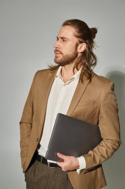 handsome and bearded businessman in beige formal attire holding laptop on grey background clipart