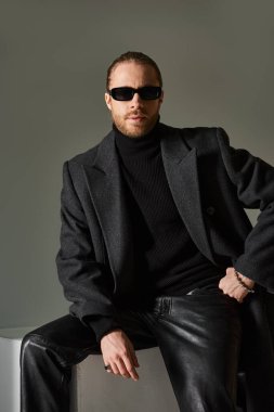 portrait of fashionable man in trendy sunglasses and coat sitting on white cube on grey backdrop clipart