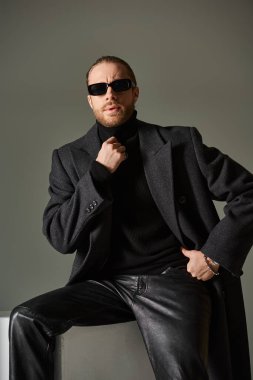 Modayı takip eden güneş gözlüklü ve gri arka planda beyaz küpte oturan moda modelin portresi