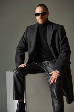 Modayı takip eden güneş gözlüklü ve gri arka planda beyaz küpte oturan moda modelin portresi